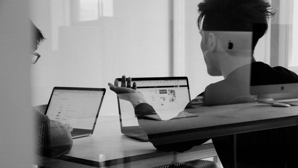 Dos personas conversando frente a notebooks sobre una mesa, vistas a través de un vidrio, en blanco y negro